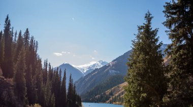 Kristal berraklığında dağ gölü, arka planda karla kaplı tepeleri olan yeşil çam ormanlarının arasına kurulmuş, parlak mavi bir gökyüzünün altında.