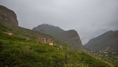 bulutlu bir gökyüzünün altında uçsuz bucaksız bir dağ manzarası. Değişen yeşil tonlarıyla yuvarlanan tepeler. Vadiden geçen dolambaçlı bir yol. Uzaktaki küçük yapılar görülebilir..