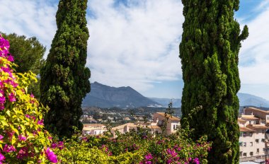 Canlı bougainvillea ve yemyeşil çerçeve bir Akdeniz kasabasının pitoresk bir manzarası. Uzun selvi ağaçları, parçalı bulutlu bir gökyüzünün altında muazzam bir zemin üzerinde dururlar..