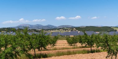 Geniş bir güneş paneli alanı, açık mavi gökyüzü altında güneşin enerjisini kullanarak araziye yayılır. Uzak dağlar bu yenilenebilir enerji çiftliğinin manzaralı güzelliğini arttırır..