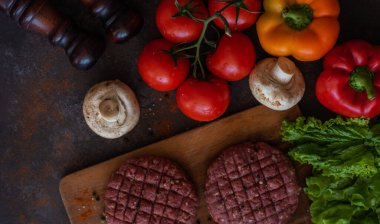 Taze doğranmış çiftlik hayvanları, siyah arka planda baharat, ot ve sebzelerle eski bir kesim tahtasının üzerinde sığır burger kızartıyorlar..