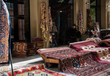 Tarihi bir kentsel bölgedeki bir dükkanın önünde renkli, karmaşık dizayn edilmiş halı ve tekstil ürünleri sergilenen bir pazar. Canlı desenler ve zengin renkler geleneksel işçiliği yansıtıyor..