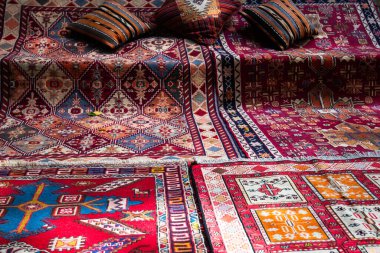 Karmaşık geometrik desenli renkli el dokuması halılar yere yayılmış ve yanında çizgili yastıklar var. Zengin renkleri, geleneksel zanaatkarlığın canlı bir gösterisini yaratıyor..