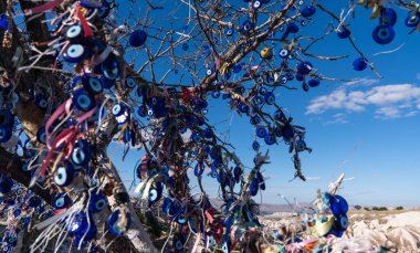 Canlı Nazar tılsımlarıyla süslenmiş, aynı zamanda Şeytani Göz olarak da bilinen bir ağaç, açık mavi gökyüzünün altında duruyor, bu da Türk kültürünün korunma ve geleneğini simgeliyor..