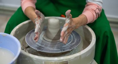 A person wearing a green apron with hands covered in clay is shaping a piece of pottery on a spinning wheel. Fully engaged in the creative process with a focus on the clay piece. clipart