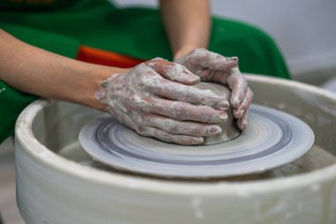 A person wearing a green apron with hands covered in clay is shaping a piece of pottery on a spinning wheel. Fully engaged in the creative process with a focus on the clay piece. clipart