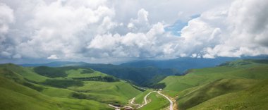 Dinamik bulutlarla dolu bir gökyüzünün altında dinamik yeşil tepelerin hareketli görüntüsü, dingin manzaranın içinden geçen dolambaçlı bir yol, yazın ilk zamanlarındaki bereketli canlılığı çağrıştırıyor..