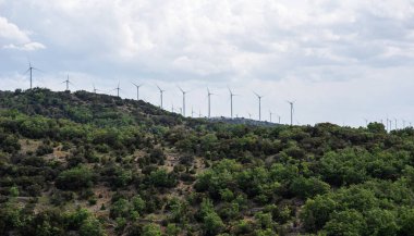 Rüzgâr türbinleri yoğun yeşillikle kaplı bir tepede bulutlu bir gökyüzünün altında dimdik durur. Manzara modern teknolojiyle doğal güzelliği, yenilenebilir enerji çabalarını harmanlıyor..