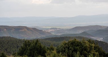 Uçsuz bucaksız tepeler ve bulutlu bir gökyüzünün altındaki sık ormanlık alanların engin manzarası, vadide yuva yapmış uzak bir köy, sakin kırsal yaşamın özünü yakalıyor..