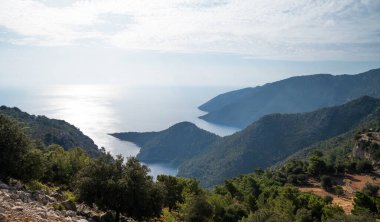 Serin bir Akdeniz kıyı şeridinin nefes kesici panoramik manzarası yemyeşil, sakin bir körfeze bakan bir yaz öğleden sonrası. Manzara, denize açılan dağlık bir bölgenin engebeli güzelliğini yakalar..