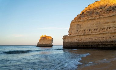 Algarve Praia da Marinha 'nın altın tepeleri batan güneşle aydınlanır. Kumlu plaja karşı sakin deniz, etkileyici kaya oluşumları çarpıcı bir kıyı manzarası yaratıyor..