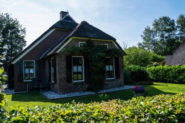 Yazın Hollanda 'nın kırsal kesimlerinde çeşitli yaprak döken ağaçlarla çevrili sık yeşil çalıların ve çiçek açan çiçeklerin arkasına kısmen saklanmış çatı evi..