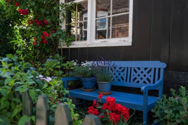 Kırmızı güller ve yemyeşil ile çevrili, parlak mavi bir bank ile bahçe köşesi, kırsal bir pencere ile kırsal siyah bir duvara karşı kurulmuş.