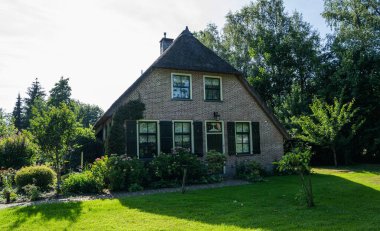 Yazın Hollanda 'nın kırsal kesimlerinde çeşitli yaprak döken ağaçlarla çevrili sık yeşil çalıların ve çiçek açan çiçeklerin arkasına kısmen saklanmış çatı evi..