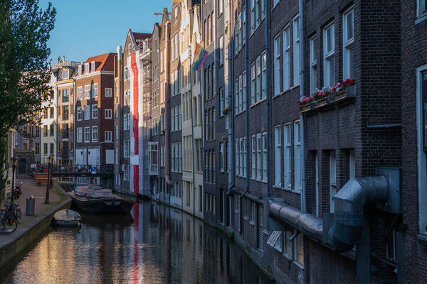 Narrow canal lined with tall brick houses in the historic center of Amsterdam, Netherlands. A moored boat, bicycles, and rainbow flag add life to the scene in soft evening light.