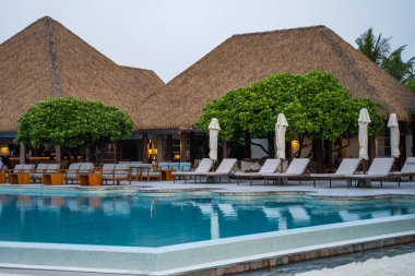 Lüks tropikal tatil köyü, sazdan çatılı bir bar, beyaz çiçekli yemyeşil ağaç, minderli ahşap şezlonglar, ve çevreyi yansıtan turkuaz yüzme havuzu.