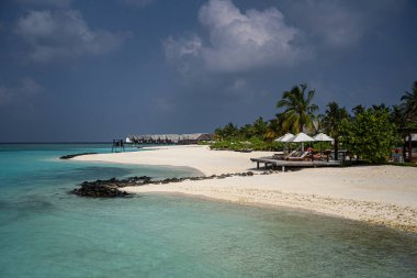 Maldivler 'in beyaz kumlu sahillerinde turkuvaz suyu, uzaktaki su villalarının üzerinde palmiye ağaçları, şemsiyeli güneş panzeleri ve dramatik bir tropik gökyüzü altında volkanik kayalar..