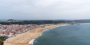 Okyanusun dalgalarının bulutlu bir gökyüzünün altında usulca kıyıları öptüğü kumlu bir sahile inen terakotta çatıları olan bir sahil kasabasının geniş bir manzarası.