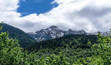 Andorra 'daki görkemli Pireneler dağları, yemyeşil ormanların üzerinde yükselen karlı tepeler. Parlak yeşillik, parçalı bulutlu bir gökyüzünün altındaki engebeli, kayalık zirvelerle çok güzel tezat oluşturuyor..