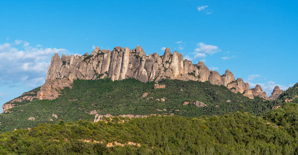 A breathtaking view of the rugged Montserrat mountain range with its distinctive rock formations, lush green forests, and clear blue sky, showcasing the natural beauty of the Catalonian landscape.