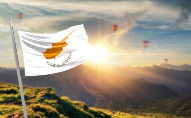 Kıbrıs bayrağı dağlarda gün batımında dalgalanıyor, arka planda sıcak hava balonları 