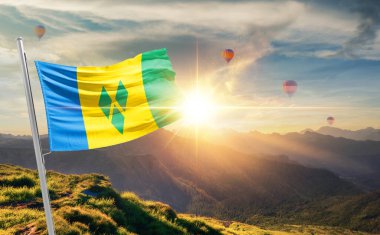 Saint Vincent ve Grenadines bayrağı dağlarda gün batımında dalgalanırken, arka planda sıcak hava balonları 