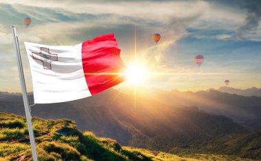 Dağlarda gün batımında dalgalanan Malta bayrağı, arka planda sıcak hava balonları 