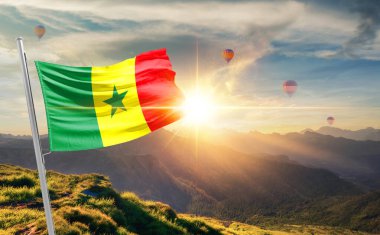 Senegal bayrağı dağlarda gün batımında sallanıyor, arka planda sıcak hava balonları. 