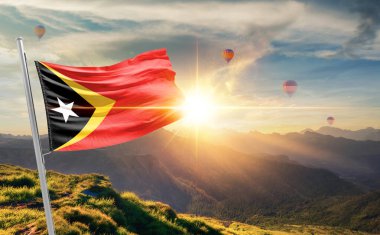 Doğu Timor bayrağı dağlarda gün batımında dalgalanıyor, arka planda sıcak hava balonları 