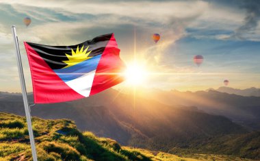 Antigua ve Barbuda bayrağı dağlarda gün batımında dalgalanırken, arka planda sıcak hava balonları 