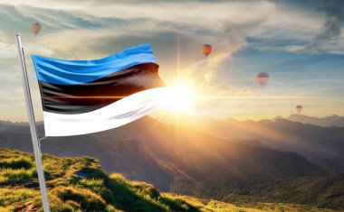 Günbatımında Estonya bayrağı sallıyor. Arka planda sıcak hava balonları var. 