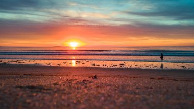 Okyanus dalgalarıyla baş döndürücü güneş ışığı altında kumlu sahilde yürüyen bir turist görüntüsü