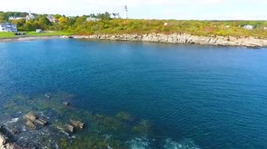 Maine 'e çarpan sığ sular ve dalgaların olduğu kayalık sahil şeridinde beyaz deniz fenerine giden Aerial' ın videosu.