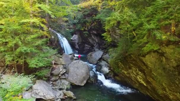緑豊かな森の岩場の渓谷の川沿いの空中ビデオ滝と岩からの滝を観察する唯一のハイカー — ストック動画