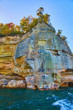 Beyaz ibikli dalgaların deniz mavisi sularında, kayalıkların altında, devrilmiş ağaçlarla çevrili görüntüsü.