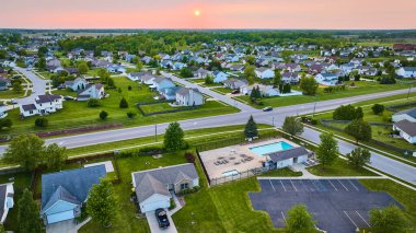 Banliyö evlerinin üzerindeki pembe günbatımı görüntüsü yeşil çimenli havuz ve otopark.