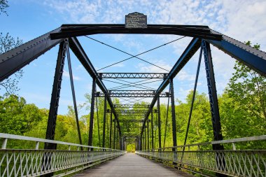 Siyah metal köprünün üzerinde beyaz bulutlar olan parlak mavi gökyüzü, yemyeşil orman arka planı
