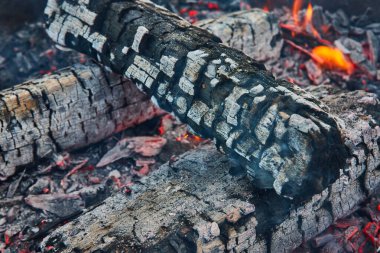 Kırık kül yüzeyi ve altında küçük turuncu alevlerle kırmızı közlü siyah kütük görüntüsü