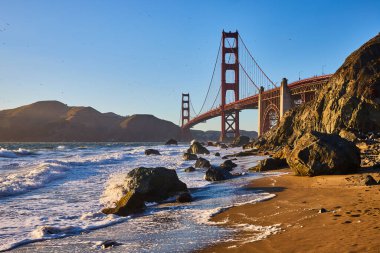 Dalgaların kumsala yuvarlanışının görüntüsü. Büyük kayalar ve gün batımına yakın Golden Gate Köprüsü.