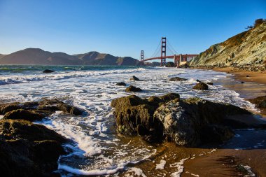 Golden Gate Köprüsü kıyısı boyunca dalgalar ve deniz köpüğüyle kumlu sahildeki yosunlu kayaların görüntüsü
