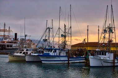 Hyde St Pier rıhtımına yanaşmış birkaç geminin görüntüsü. Arkalarında günbatımı altınları ve dağın üzerinde puslu bulutlar var.