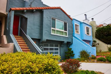 Kırmızı kiremit çatılı mavi renkli bir ev ve mavi merdivenli parlak kırmızı bir kapı.