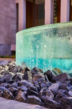 Binanın önündeki büyük siyah kayaların üzerine düşen suyun çarpık camlı dairesel duvar görüntüsü