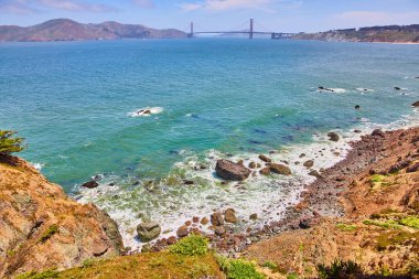 Dalgalar ve kayaların etrafında dönen deniz köpüğü ve uzak Golden Gate Köprüsü ile Rocky sahili görüntüsü