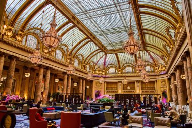 Parlak tavanın altında altın turuncu avizeler ve sandalyeli insanların olduğu abartılı bir salon.