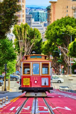 Tepeye doğru yükselen kırmızı ve bronz tramvay görüntüsü. Arkasında güneşli yeşil çınar ağaçları var. San Francisco, CA