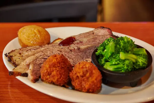 Hearty Southern Cuisine: Indulge in a mouthwatering plate of smoky brisket, glossy barbecue sauce, crispy hush puppies, golden croquettes, and vibrant broccoli. A delicious snapshot of comfort food at