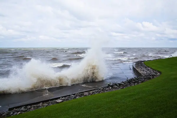 Dynamic clash of nature and structure as dark waves crash against seawall in overcast weather, creating a striking contrast with serene green lawn. Perfect for illustrating environmental challenges