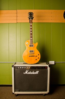 Rock 'n Roll Vibes: Bir güneş patlaması elektro gitarı, keskin bir zıtlıkla yeşil bir duvara yaslanır ve parlak, parlak vücuduna dikkat çeker. İkonik Marshall amplifikatörü hazır.