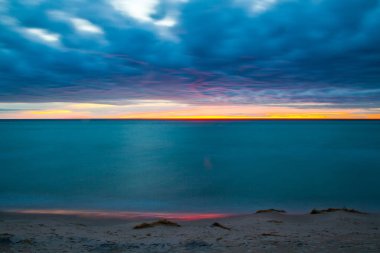 Michigan Gölü üzerinde sakin bir günbatımı, cam gibi sulara yansıyan canlı renkler. Dokunulmamış kum ve bitki örtüsüyle çerçevelenmiş sakin bir sahil rüya gibi, ruhani ortamı güçlendirir. Tam oldu.